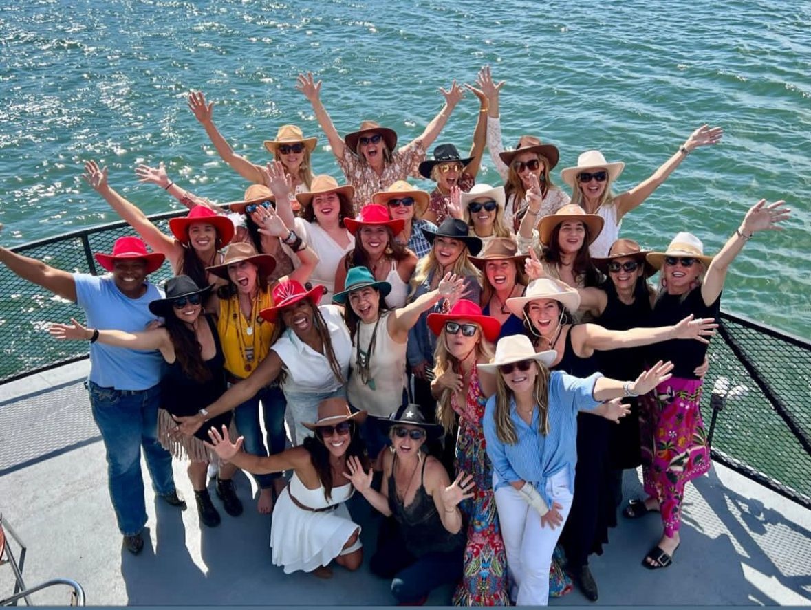 Group of people in colorful outfits and hats posing on a boat with arms raised on the water