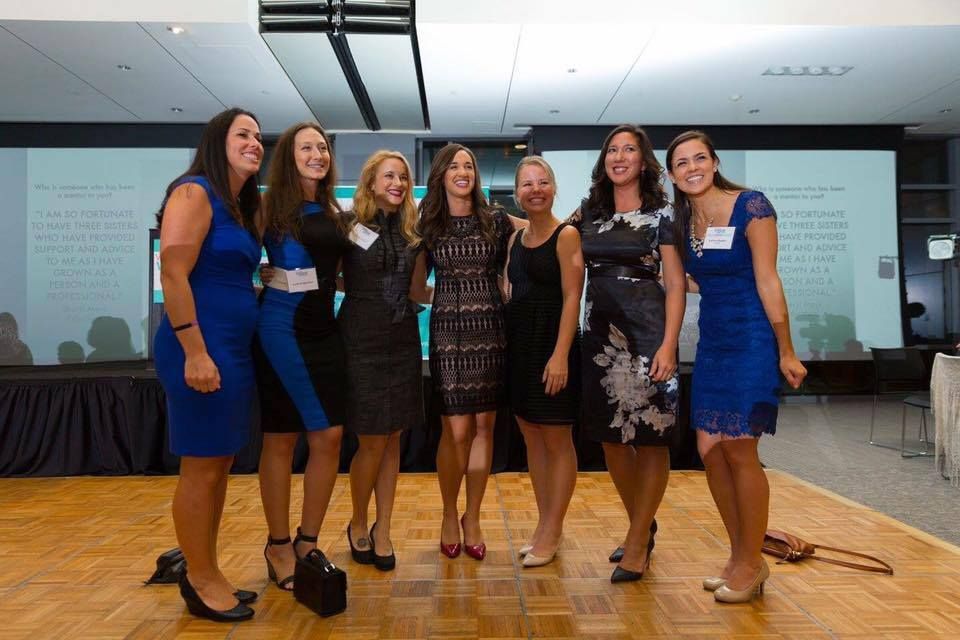 Seven women pose smiling in dresses on a wooden floor at an indoor event.