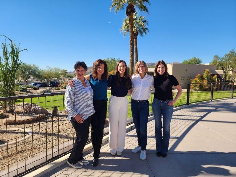 Five people standing outdoors on a sunny path beside a fence and palm tree, smiling at the camera.